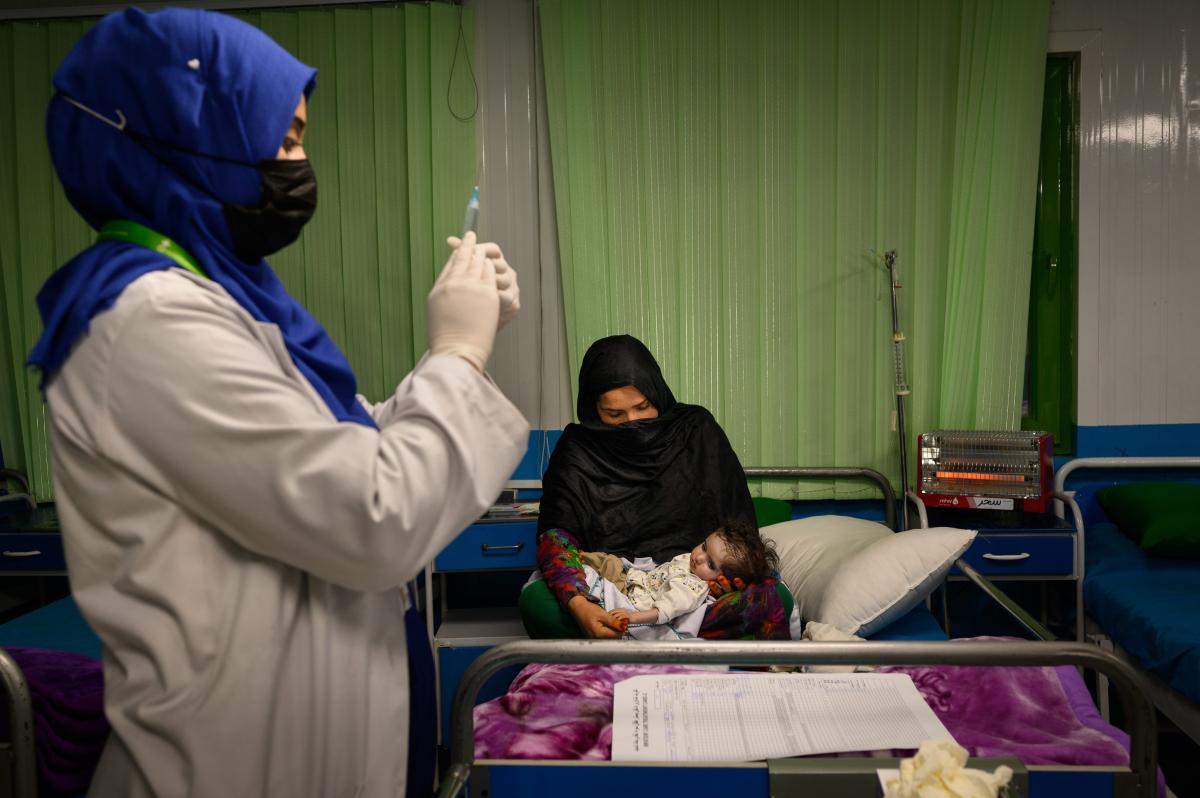 Una mujer afgana sentada en su cama con su nieta de un mes y medio, que padece desnutrición aguda grave y neumonía, mientras una enfermera se prepara para administrar a la niña medicación intravenosa en una sala de hospitalización de la unidad de alimentación terapéutica de Acción contra el Hambre 